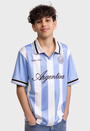 Fiatal fiú göndör hajjal, kék-fehér csíkos argentin focimezt viselve, zsebre tett kézzel egy egyszerű háttér előtt áll.