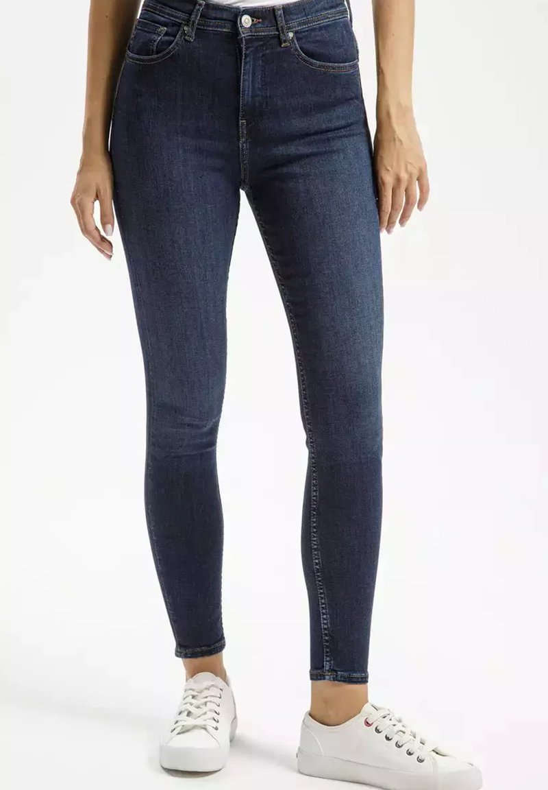 Ciemnoniebieskie wąskie dżinsy z wysokim stanem, pięcioma kieszeniami i gładką fakturą dżinsu, w połączeniu z białymi sneakersami.