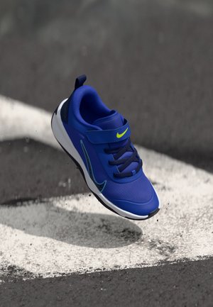 Scarpa sportiva blu con tomaia in rete, dettagli neon, cinturino regolabile e suola leggera, mostrata sospesa sopra una linea bianca su un marciapiede.