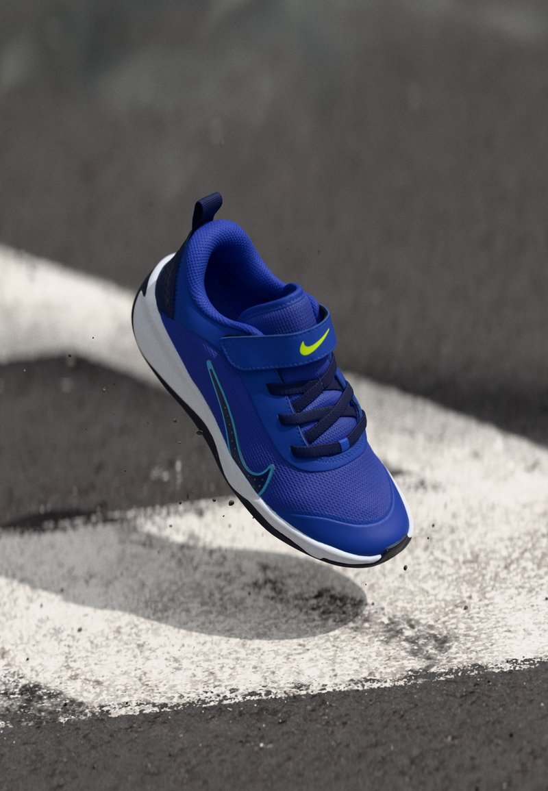 Zapatilla deportiva azul con parte superior de malla, acentos neón, correa ajustable y suela ligera, mostrada flotando sobre una línea blanca en el pavimento.