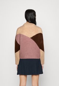 Femme portant un pull à blocs de couleur beige, rose et marron avec des manches longues et une jupe plissée sombre, vue de dos.