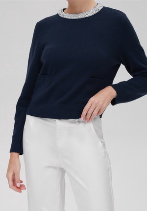 Femme portant un pull bleu marine avec un col orné de perles et un pantalon blanc, posant avec une main sur la hanche et l'autre détendue le long du corps.