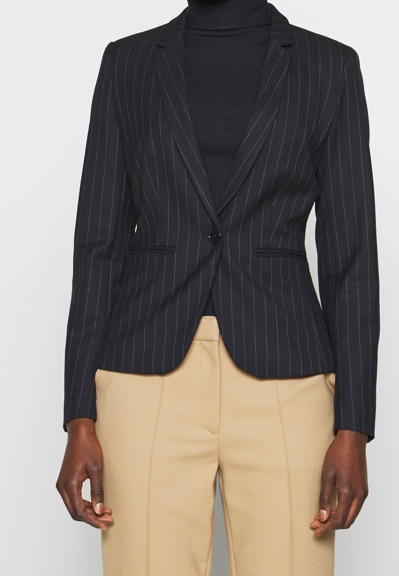 Czarna marynarka w prążki z jednym guzikiem noszona na czarnym golfie, w połączeniu z beżowymi spodniami od garnituru.