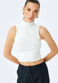 White ribbed sleeveless top with a mock neck, cropped length, and textured fabric. Paired with dark pinstriped trousers.