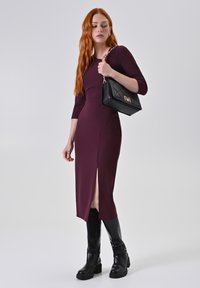 Bordeaux gekleed met driekwart mouwen en een zijsplit, gecombineerd met een zwarte gewatteerde handtas en zwarte laarzen tot de knie.
