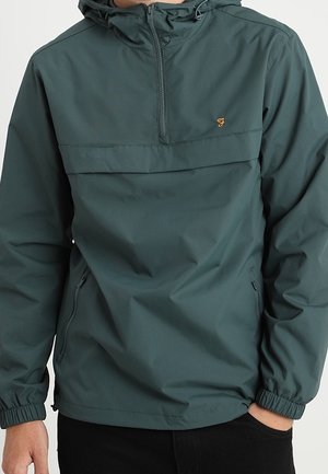 Hombre con chaqueta cortavientos con capucha de color verde oscuro, media cremallera y bolsillos laterales con cremallera, mano en el bolsillo, sin marcas visibles.