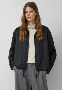 Młoda kobieta z długimi falującymi włosami w czarnej zapinanej na guziki kurtce na beżowym golfie i szarych spodniach w cienkie prążki, z rękami w kieszeniach.