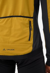 Heren overhemd met lange mouwen van gele stof met zwarte accenten, een zijritszak en logodetail nabij de zoom.