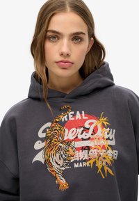 Hoodie gris avec un design de tigre brodé en orange et noir, avec un texte graphique et des accents de palmier en jaune et blanc.