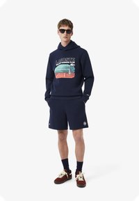 Mörk marinblå huvtröja med grafisk tryck, parat med matchande shorts. Accenteras av röda och vita skor samt svarta solglasögon.