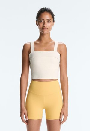 Nő áll előre nézve, krém színű rövidített topban és magas derekú sárga biciklis rövidnadrágban, egyszínű háttér előtt.