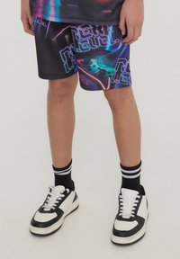 Pantaloni corti neri con un design grafico colorato che presenta elementi di gioco e il testo "Prossimo Livello". Abbinati a sneakers bianche e nere e calzini a righe.