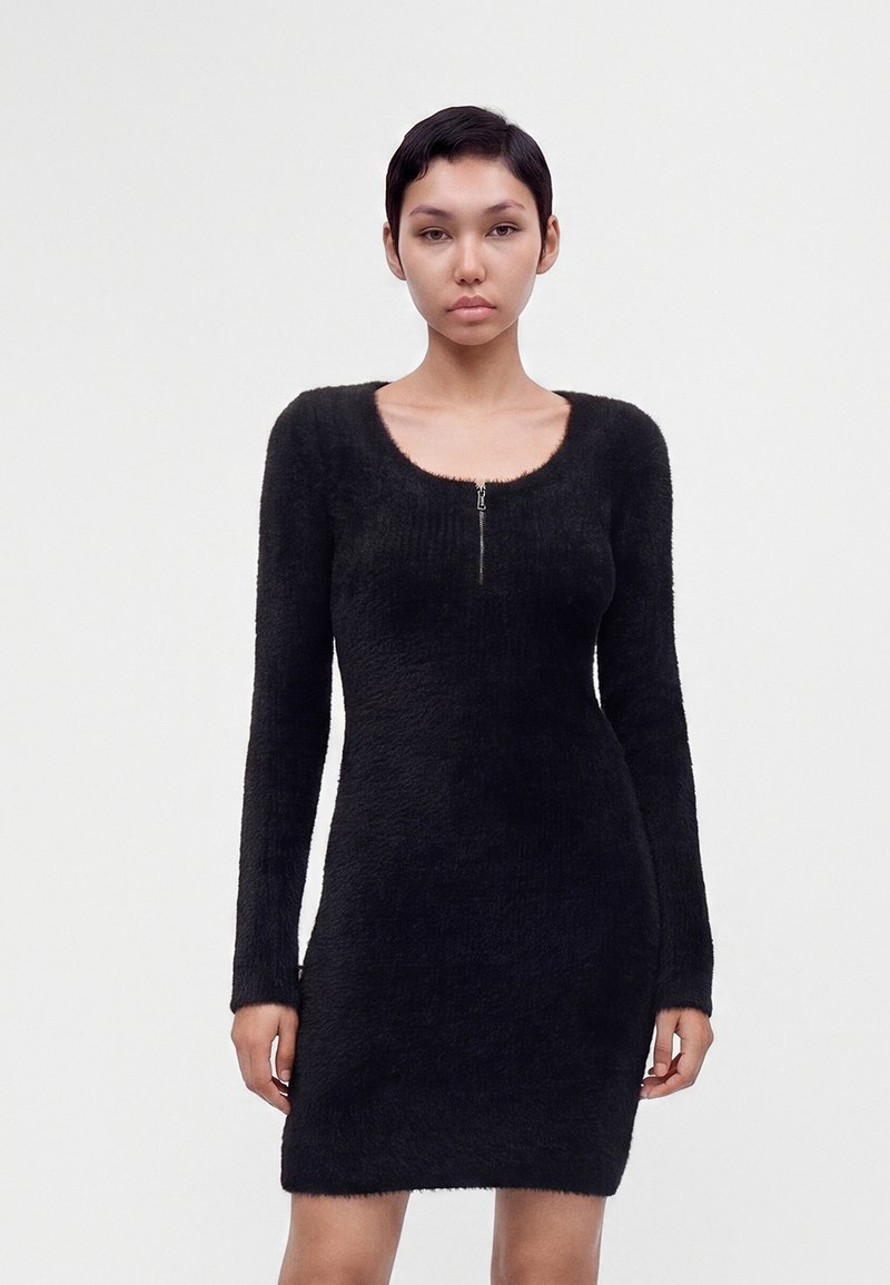 Femme aux cheveux courts portant une robe noire duveteuse à manches longues avec une fermeture éclair à l'avant, debout devant un fond blanc uni.