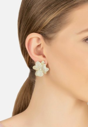 Gold flower-shaped earrings feature a textured surface with small sparkling accents and a central rhinestone, worn on an ear.