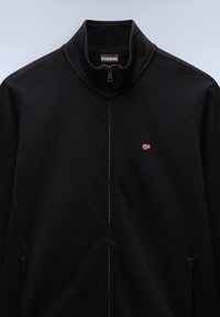 Schwarze Zip-Jacke mit hohem Kragen, ausgestattet mit einem kleinen rot-weißen bestickten Logo auf der Brust. Glattes Material, minimales Design.