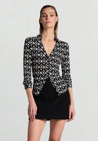 Schwarz-weiß gemusterte Bluse mit Diamantmuster, langen Ärmeln, kragenlosem Ausschnitt und figurbetonter Silhouette.