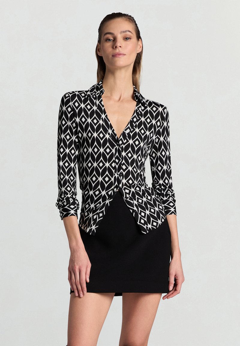 Schwarz-weiß gemusterte Bluse mit Diamantmuster, langen Ärmeln, kragenlosem Ausschnitt und figurbetonter Silhouette.