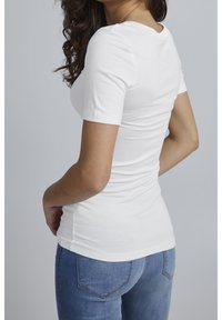 Femme aux cheveux bruns bouclés portant un t-shirt blanc uni ajusté à manches courtes et un jean en denim bleu, debout devant un fond gris.