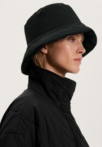 Kvinna i svart vadderad jacka med hög krage och svart bucket hat, vänd åt höger mot neutral bakgrund.