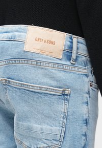 Τζιν παντελόνι ανοιχτού μπλε χρώματος με δερμάτινη ετικέτα που γράφει "ONLY & SONS." Ορατές ραφές, δύο πίσω τσέπες και ξεθωριασμένη υφή.