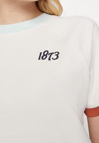 Bílá tričko s krátkými raglánovými rukávy, světle modrým límcem a oranžovými manžetami. Obsahuje vyšitý nápis "1873" v tmavě modré. Jemná textilie.