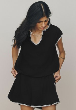 Frau mit langen dunklen Haaren, die ein schwarzes Top und einen Rock mit weißem Zickzackbesatz trägt, schaut nach unten, Hände in den Taschen vor einfachem Hintergrund.