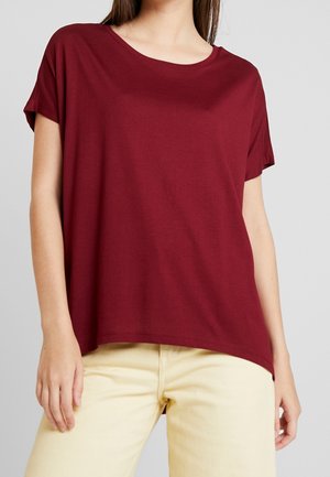 Vrouw draagt een losse, bordeauxrode top met korte mouwen, gecombineerd met lichtbeige broek, staand tegen een effen grijze achtergrond.