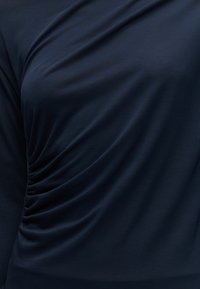 Detailní záběr námořnicky modré látky se shrnutím na boku, ukazující texturu a pružnost materiálu.