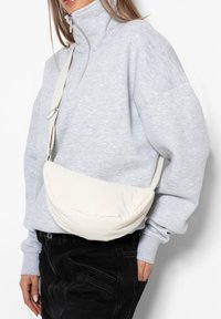 Woman wearing a light gray zip-up sweatshirt and black pants with a cream-colored padded crossbody bag across her torso.