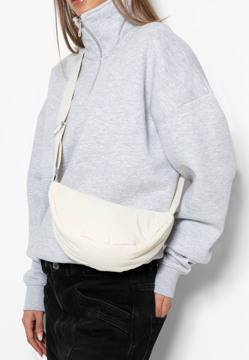 Woman wearing a light gray zip-up sweatshirt and black pants with a cream-colored padded crossbody bag across her torso.