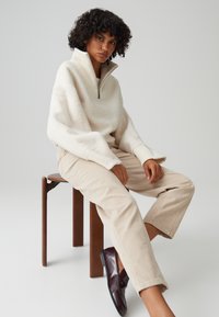 Pullover in pile color crema con colletto alto e zip, abbinato a pantaloni in velluto a coste beige. Scarpe bordeaux completano il look. Seduto su uno sgabello di legno.