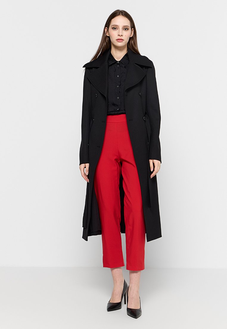 Longo cappotto nero con un design su misura, indossato sopra una camicia nera e pantaloni rossi a vita alta. Tacchi alti neri completano l'outfit.