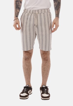 Man wearing beige and black vertical striped shorts with white drawstring, white t-shirt, black and white sneakers, and arm tattoos visible.