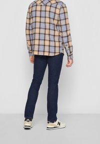 Camicia di flanella a quadri nei toni beige, blu e marrone, abbinata a jeans di denim scuro. Le scarpe sono chiare con accenti scuri e un logo.