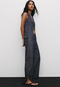 Jeansväst med öppen front och smal krage, i kombination med vida denimbyxor med sidoklipp och knytningar i mörkblått.