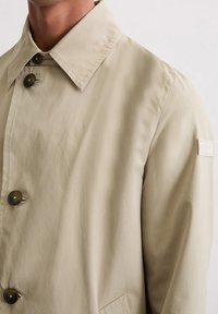 Beige trenchcoat met een puntige kraag, twee grote bruine knopen en een gestikt logo op de schouder. Soepele, lichte stoftextuur.