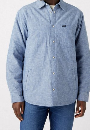 Man wearing light blue button-up shirt with snap buttons, chest pocket with Wrangler logo, and blue jeans, standing against white background.