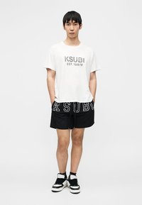 Λευκό βαμβακερό t-shirt με εκτύπωση 'KSUBI EST. 1999', συνδυασμένο με μαύρο σορτς με έντονα λευκά γράμματα και μαύρα και λευκά αθλητικά παπούτσια.