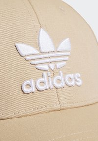 Gorra de béisbol marrón hecha de una mezcla de algodón, con un logo de Adidas bordado en blanco y un diseño de tres franjas en la parte frontal. Textura suave.