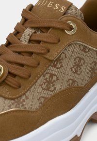 Baskets marron et beige avec le logo "Guess" sur la languette, côtés en tissu à motifs, lacets marron et semelle blanche avec des accents marron.