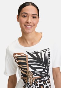 Weißes T-Shirt mit kurzen Ärmeln, das mit Tierdruck und Blattdesigns in Schwarz und Braun verziert ist, zusätzlich mit strukturellen Elementen wie Punkten.