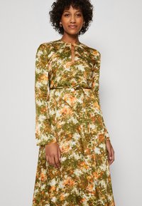 Robe à manches longues avec un motif floral aux teintes vertes, orange et blanches, présentant un décolleté en forme de clé et une ceinture en tissu à la taille.