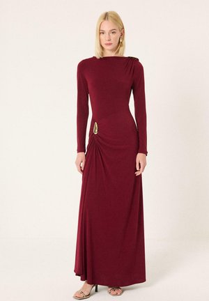 Γυναίκα με ξανθά μαλλιά φορώντας μακρυμάνικο, μακρύ φόρεμα μπορντό με χρυσές λεπτομέρειες από καρφίτσα, που στέκεται μπροστά σε απλό ανοιχτό φόντο.