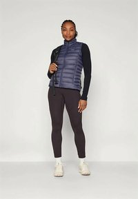 Jūrinio mėlynumo pūkų liemenė su užtrauktuku priekyje, juodi ilgomis rankovėmis marškiniai, derinami su tamsiomis leginsais ir šviesiais sportiniais batais. Laikydama žygių lazdą.