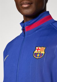 Blå zip-jakke med rød og blå stribe, der har et broderet FC Barcelona-logo; glat tekstur, sporty design.