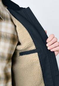 Chaqueta negra con un suave forro de forro polar beige, que muestra un exterior liso y un detalle de bolsillo en el lado izquierdo del forro.