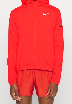 Personne portant une veste zippée Nike rouge vif avec capuche et un short de sport rouge assorti sur un fond uni.