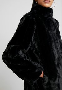 Svart faux fur-kappa med hög krage, överdimensionerade ärmar och sidofickor. Mjuk textur med en rik, djup svart färg.