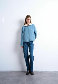 Jeune femme debout à l'intérieur, portant une veste polaire bleu clair avec un col blanc, un jean bleu et des bottines marron foncé.