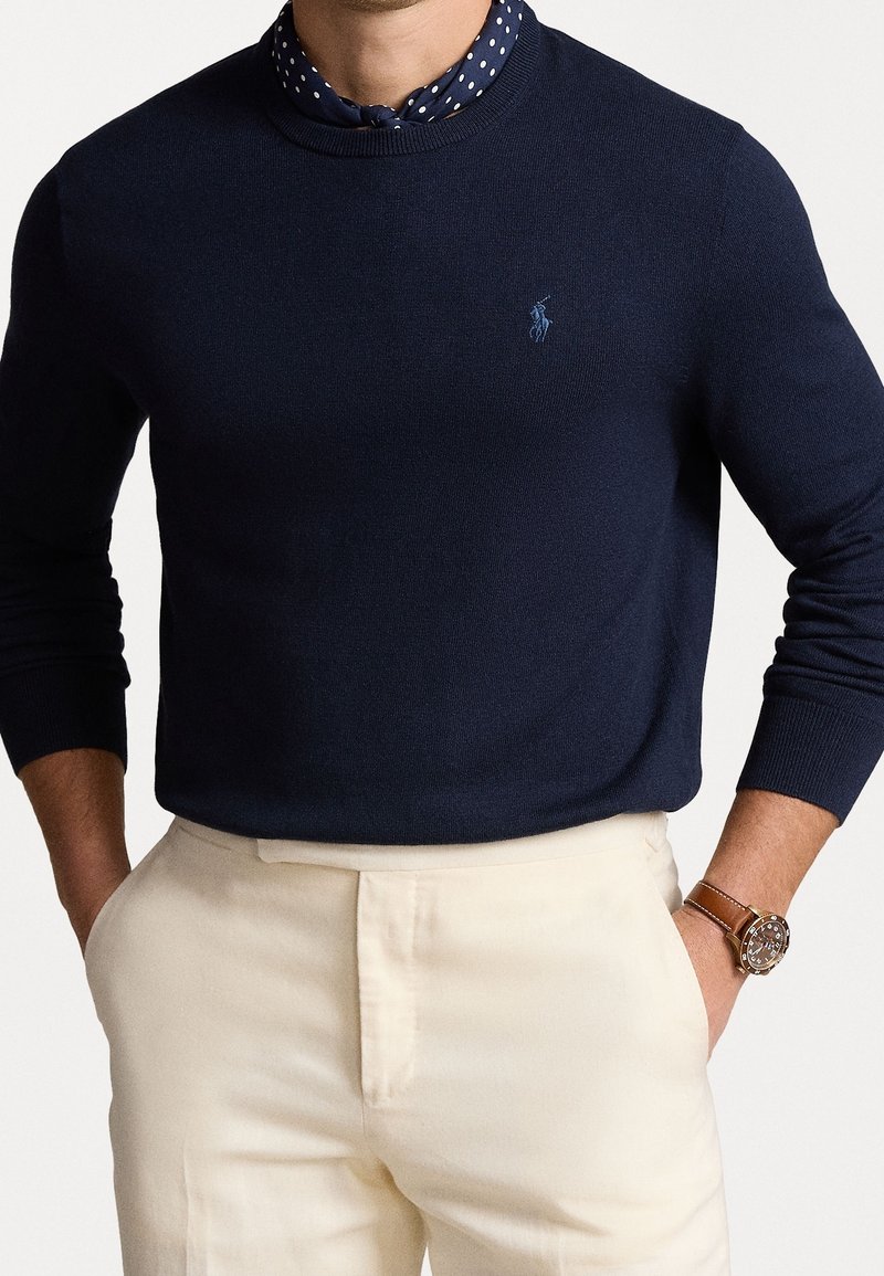 Pull en tricot bleu marine avec col rond et poignets côtelés, porté par-dessus une chemise à col bleu marine à pois. Pantalon habillé crème et montre marron inclus.